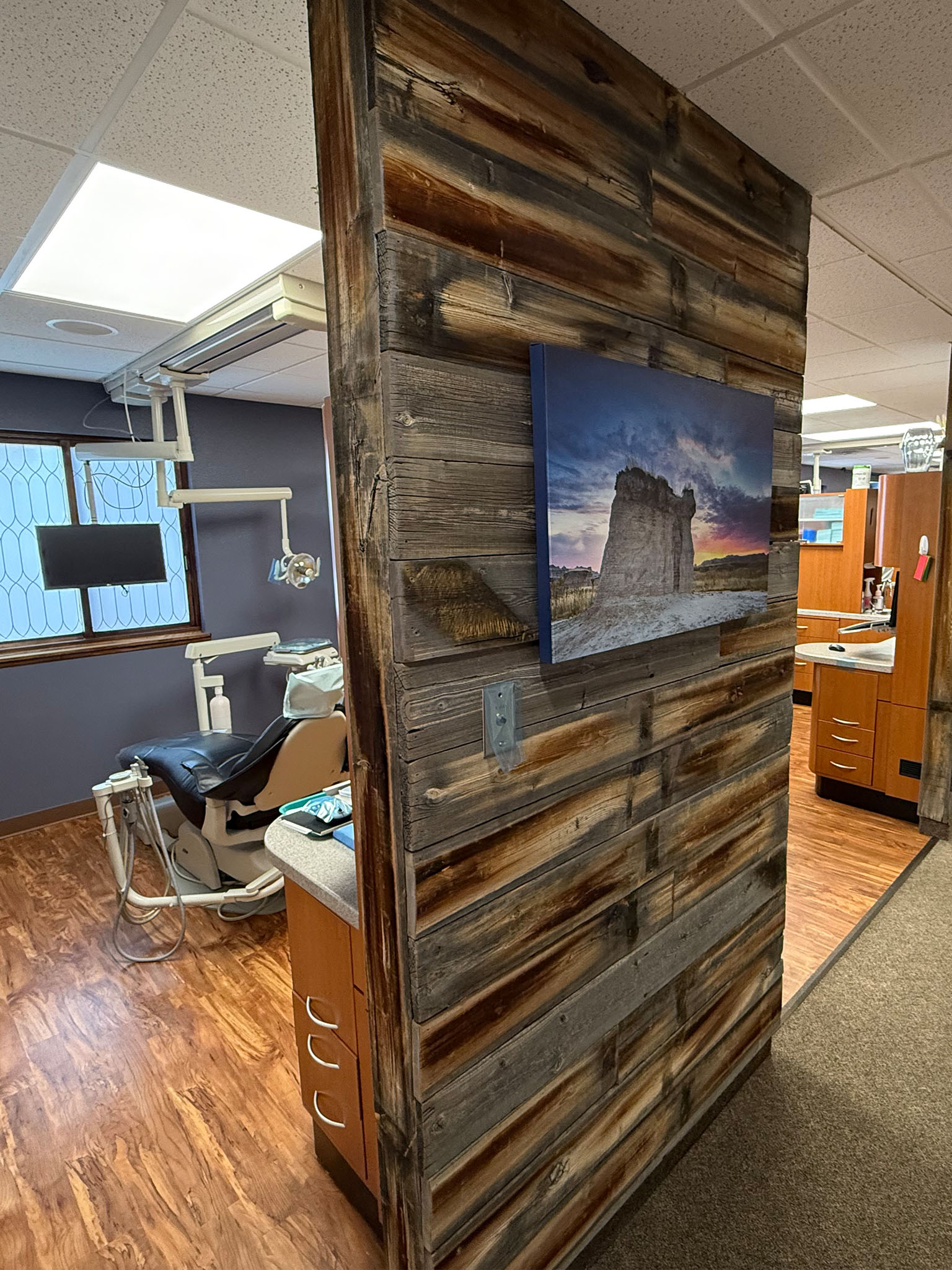The image depicts a dental office with a rustic wall decoration featuring a framed photograph and a wooden plank texture, set against a backdrop of medical equipment and a window with natural light.