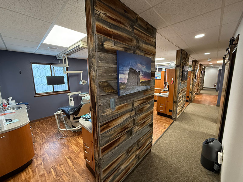 The image shows an interior view of a dental office with a rustic decor theme, featuring wood paneling, a large window, dental equipment, and a waiting area.