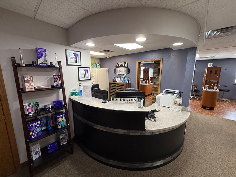 The image depicts an interior view of a well-lit office reception area with a curved counter, a desk with a computer monitor and keyboard, a bookshelf with various items, and a wall displaying framed certificates and awards.