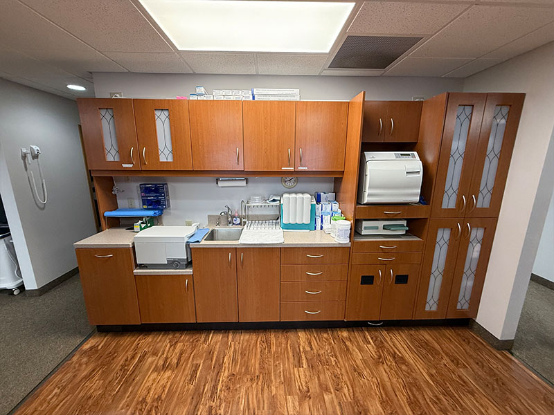 The image shows an interior view of a dental office with wooden cabinets, a countertop with a sink, various medical equipment, and a large window allowing natural light into the room.