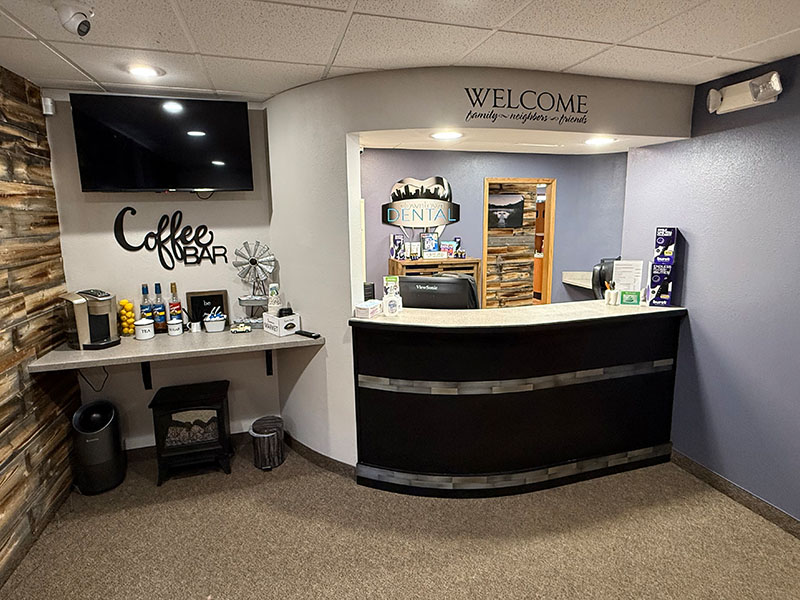 The image shows an interior view of a coffee shop with a welcoming sign, a counter with a cash register and coffee-making equipment, and a cozy seating area.