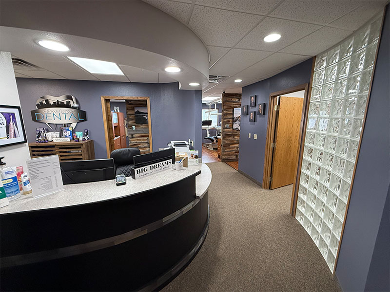 The image depicts an interior view of a dental office reception area with a modern design, featuring a desk with a computer monitor and keyboard, a glass wall displaying various items, and a large window allowing natural light into the space.